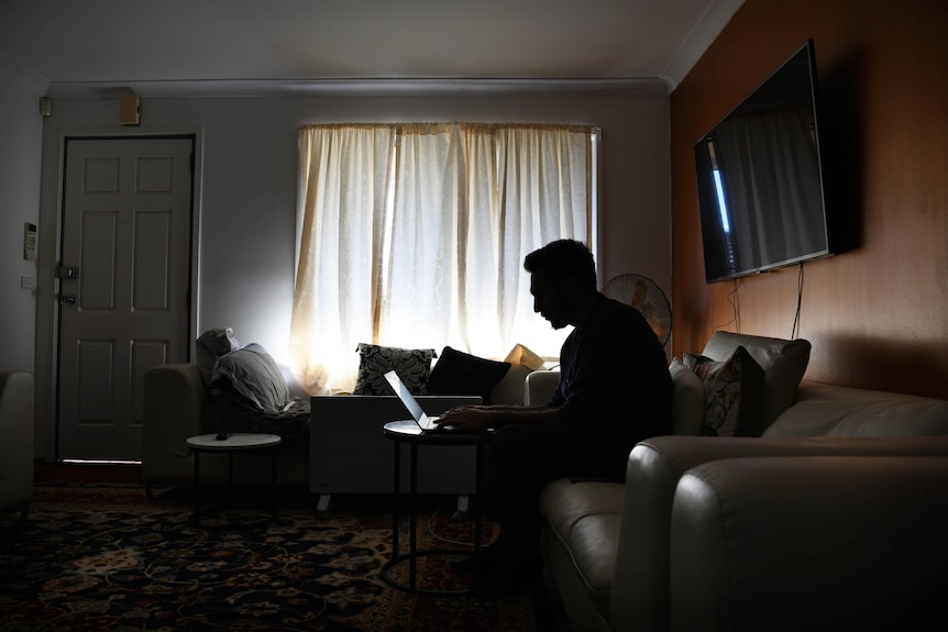 A man in silhouette sitting in a lounge room on a counch, leaning over his laptop
