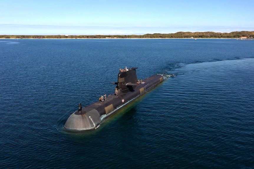 A submarine moves through the water towards the camera, in the day time. The shoreline can be seen in the distance.