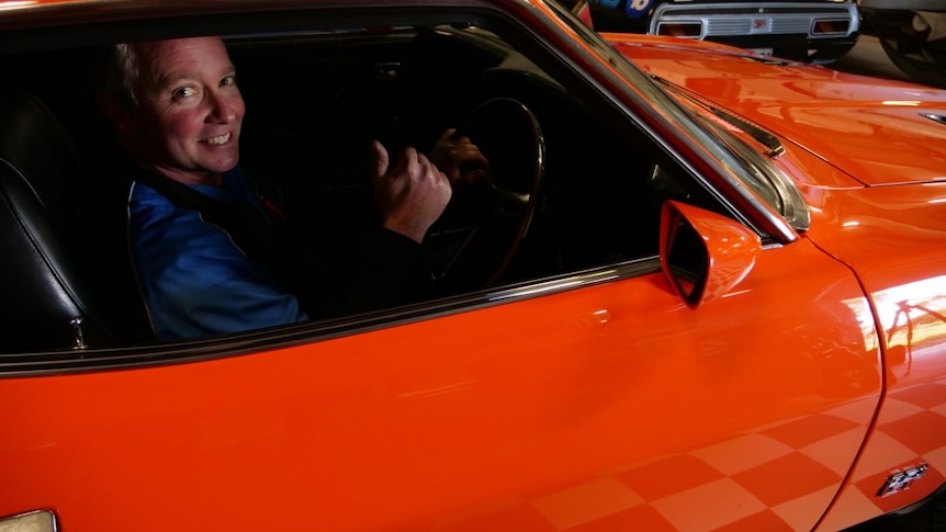 A grinning man smiles with his thumbs up while sitting in a bright orange Ford Falcon