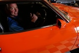 A grinning man smiles with his thumbs up while sitting in a bright orange Ford Falcon