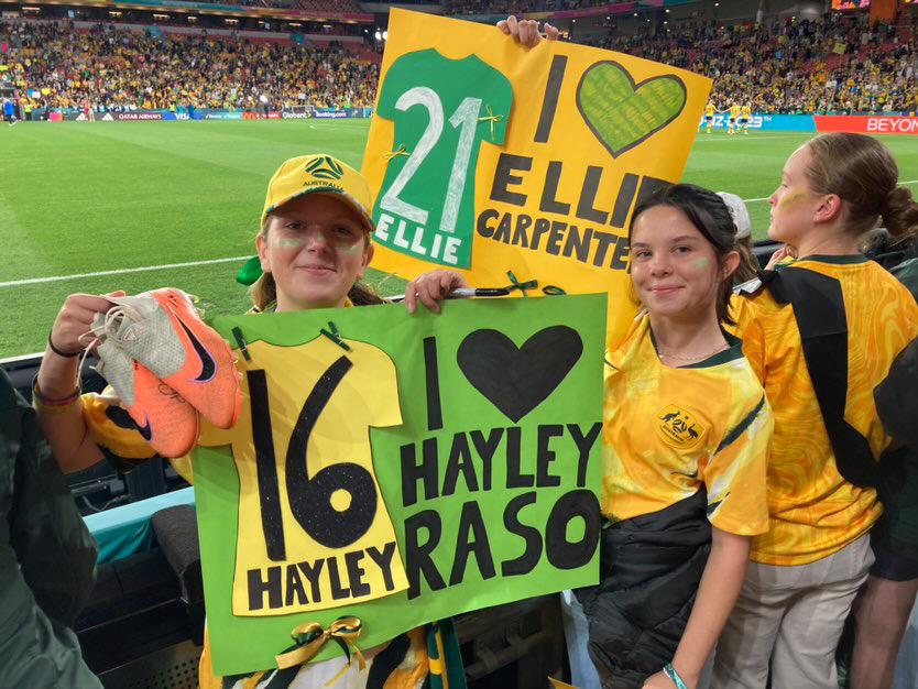 Two teens in Australian sporting colours in the stands at a stadium, holding a pair of boots and a poster.