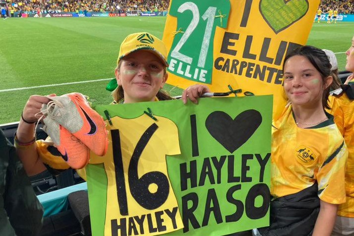 two teenage girls at stadium in green and gold holding posters and a pair of soccer boots