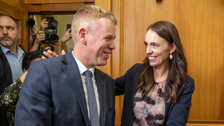 A smiling woman goes to hug a smiling man as they stand in front of a wood panelled wall.