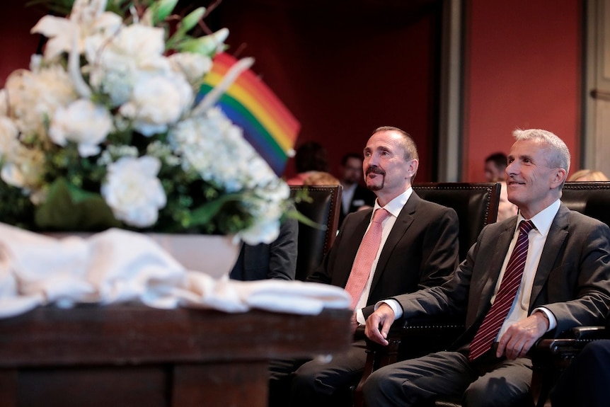 Karl Kreile and Bodo Mende sit in a chair next to each other behind a bouquet of white flowers with a rainbow flag.
