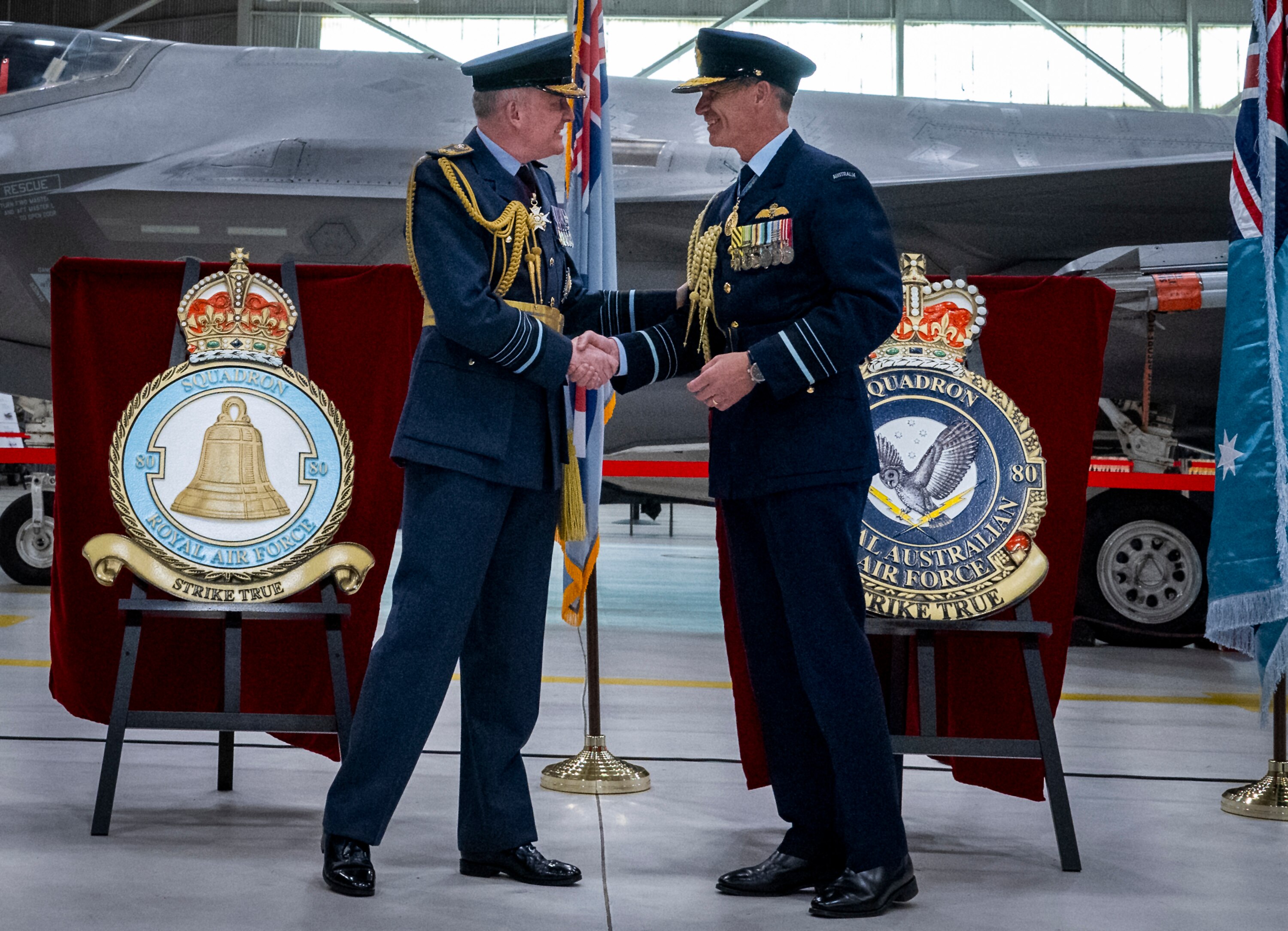 Two men in military dress shake hands in an air hangar, podiums with military emblems are behind them.