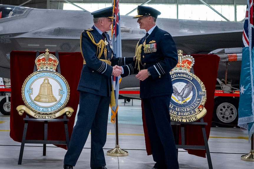 Two men in military dress shake hands in an air hangar, podiums with military emblems are behind them.