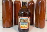 A brown glass bottle of EZY Sauce, against 4 tall bottles of tomato sauce