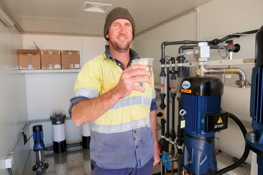 A man wearing a high vis work shirt and a beanie stands next to a water desalination unit.