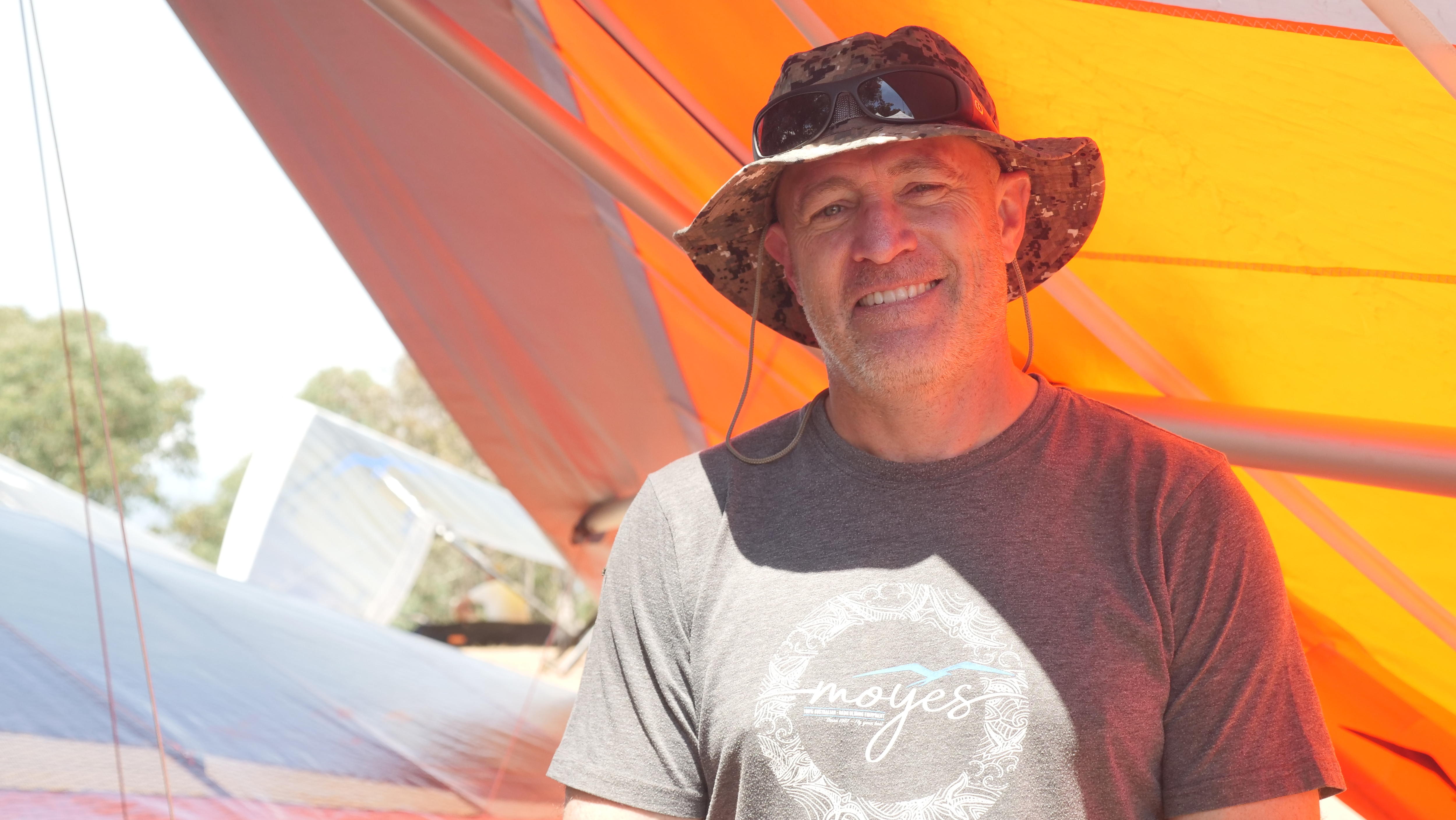 Um homem sob uma asa delta laranja sorri para a câmera. 