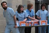 In front of a sandstone building, four people carry a stretcher with four orange boxes that read 'save our NHS'