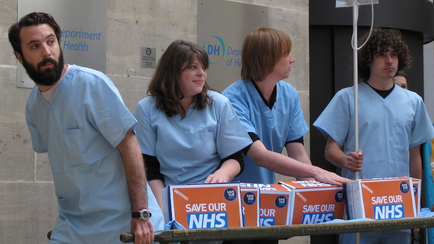 In front of a sandstone building, four people carry a stretcher with four orange boxes that read 'save our NHS'