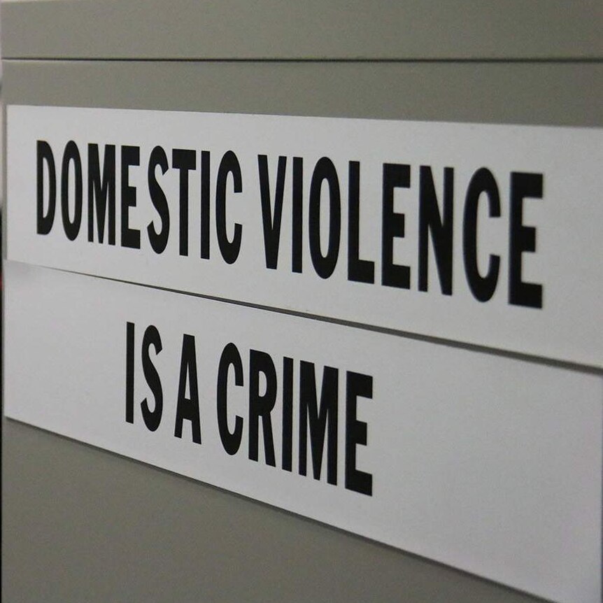 A sign saying 'domestic violence is a crime' on a filing cabinet, with a Queensland police officer in background.
