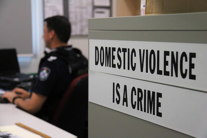 A sign saying 'domestic violence is a crime' on a filing cabinet, with a Queensland police officer in background.