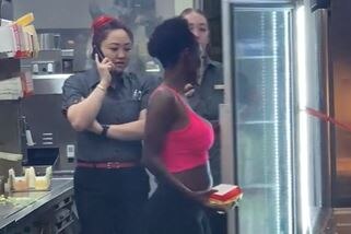 A woman wearing a bright pink top faces a fridge, with a burger box in one hand. Two staff members are behind her