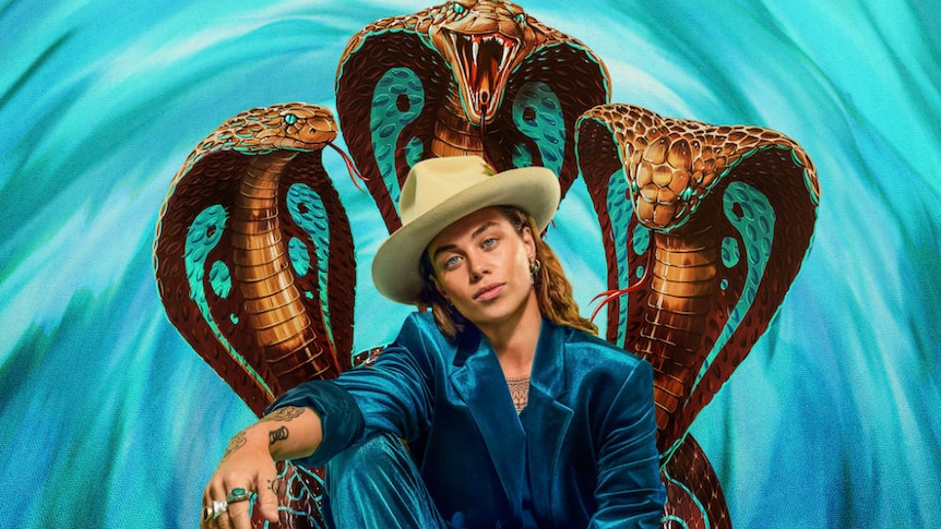 Tash Sultana in blue suit, wearing beige hat,  three cobra snakes behind them