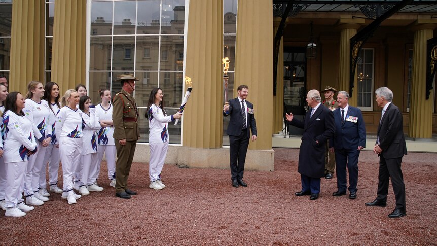 King Charles and his staff stand right, two people hold torches aflame centre, and a group of torchbearers in white left