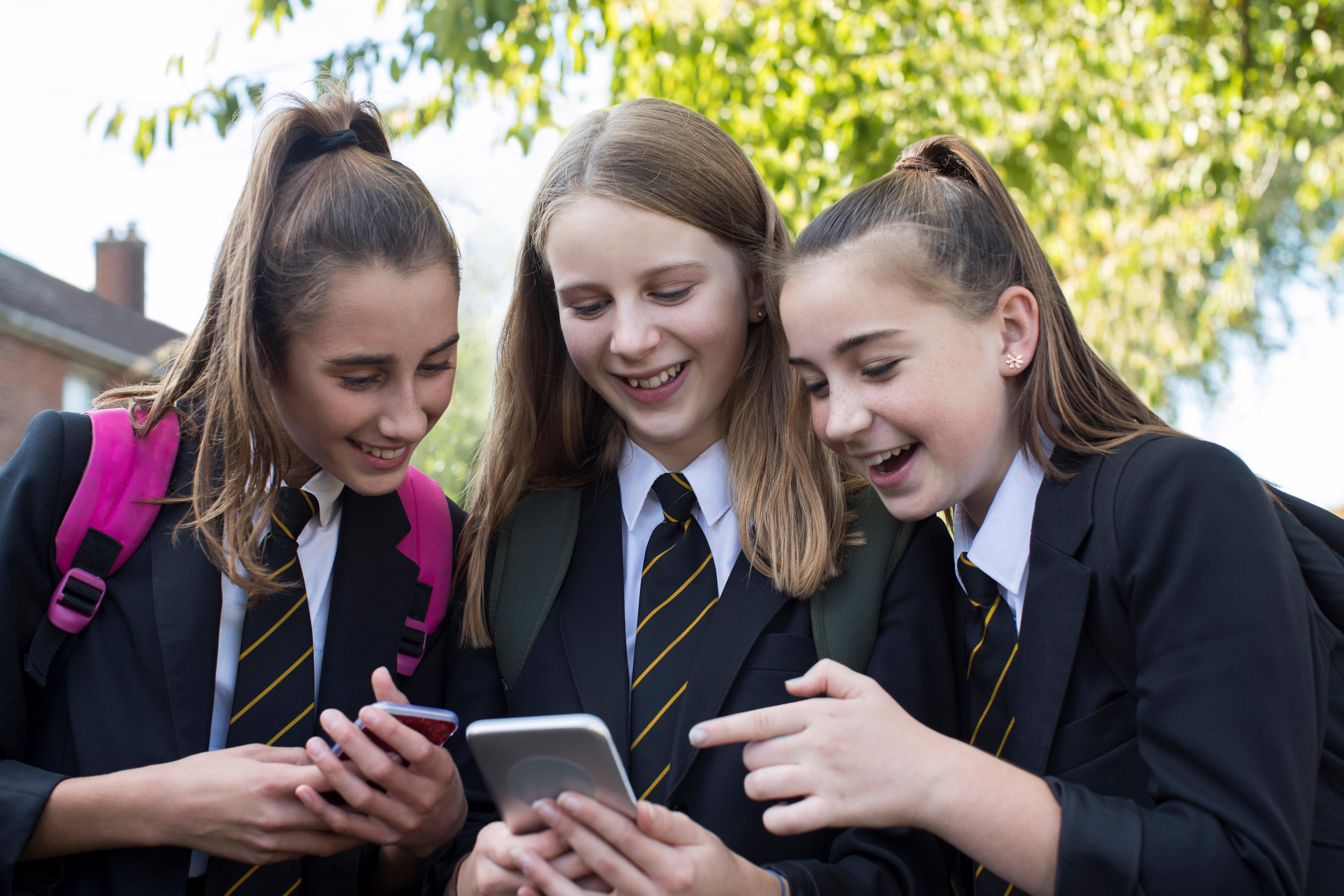 Three smiling female high school students in uniform look at their mobile phones outside of school.