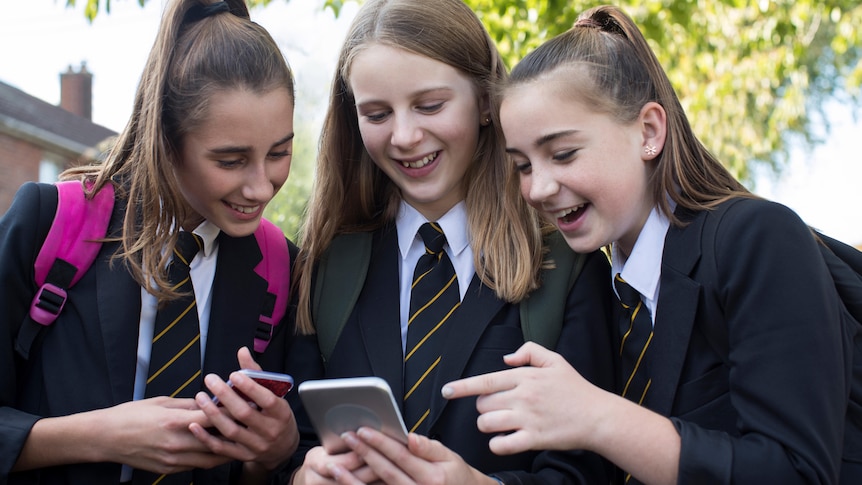 Three smiling female high school students in uniform look at their mobile phones outside of school.