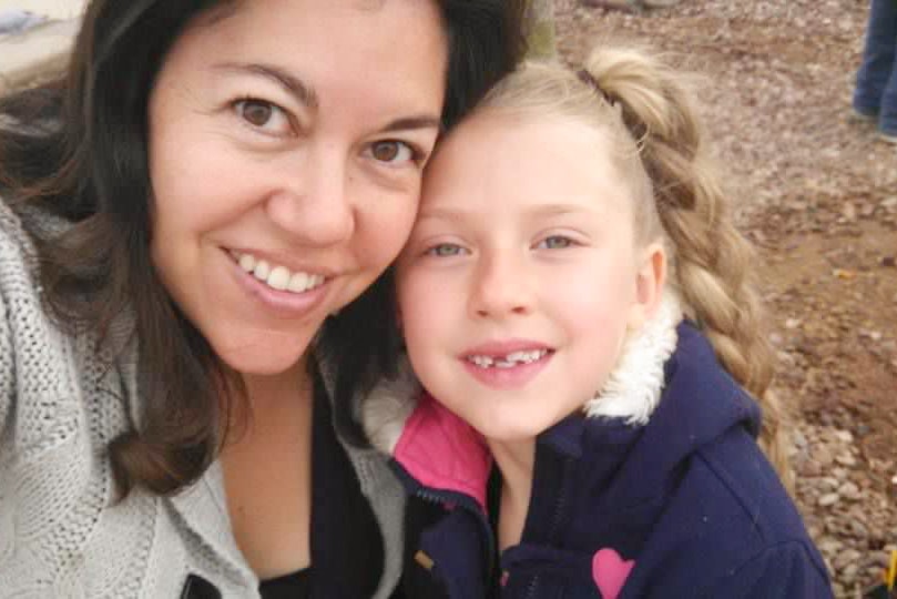 A woman with dark hair and eyes smiles next to a girl with blue eyes and blonde hair.