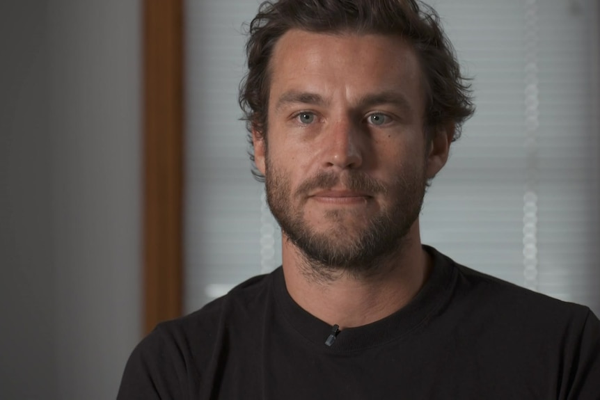 A young man with a beard is interviewed for television in a black t-shirt. He is looking serious.