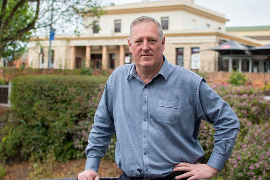 A middle-aged man standing in gardens with hand on hip, half smile, a stately cream house with pillars behind him.