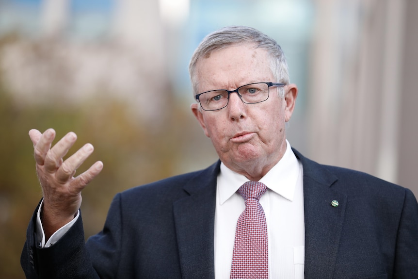 Coulton is outdoors, mid speech, right arm raised, wearing dark blue suit and glasses.