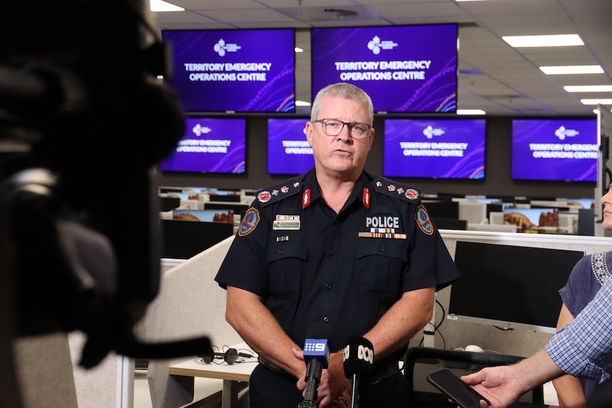 A man wearing a police uniform standing in front of TV screens in an office building