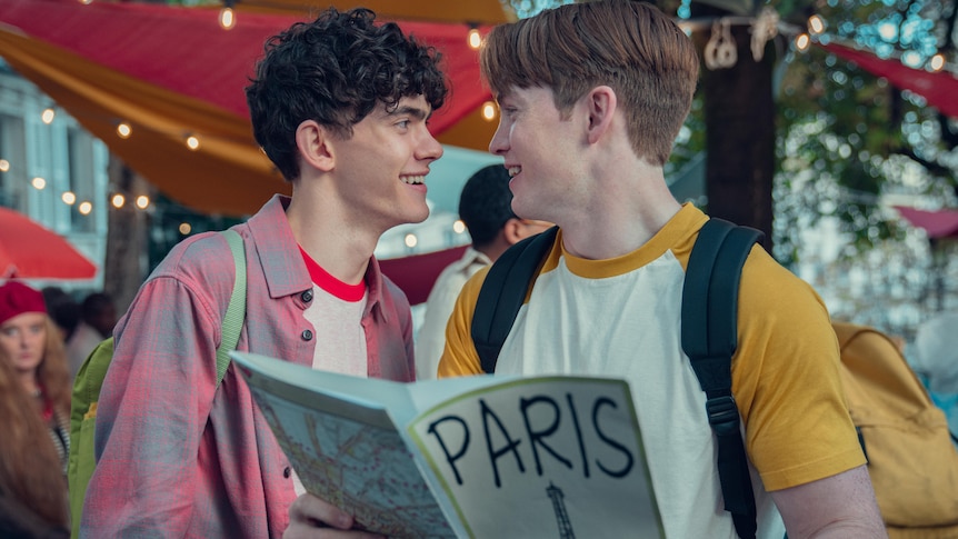The characters Nick and Charlie from Heartstopper hold open a Paris guidebook and look into each other's faces with excitement