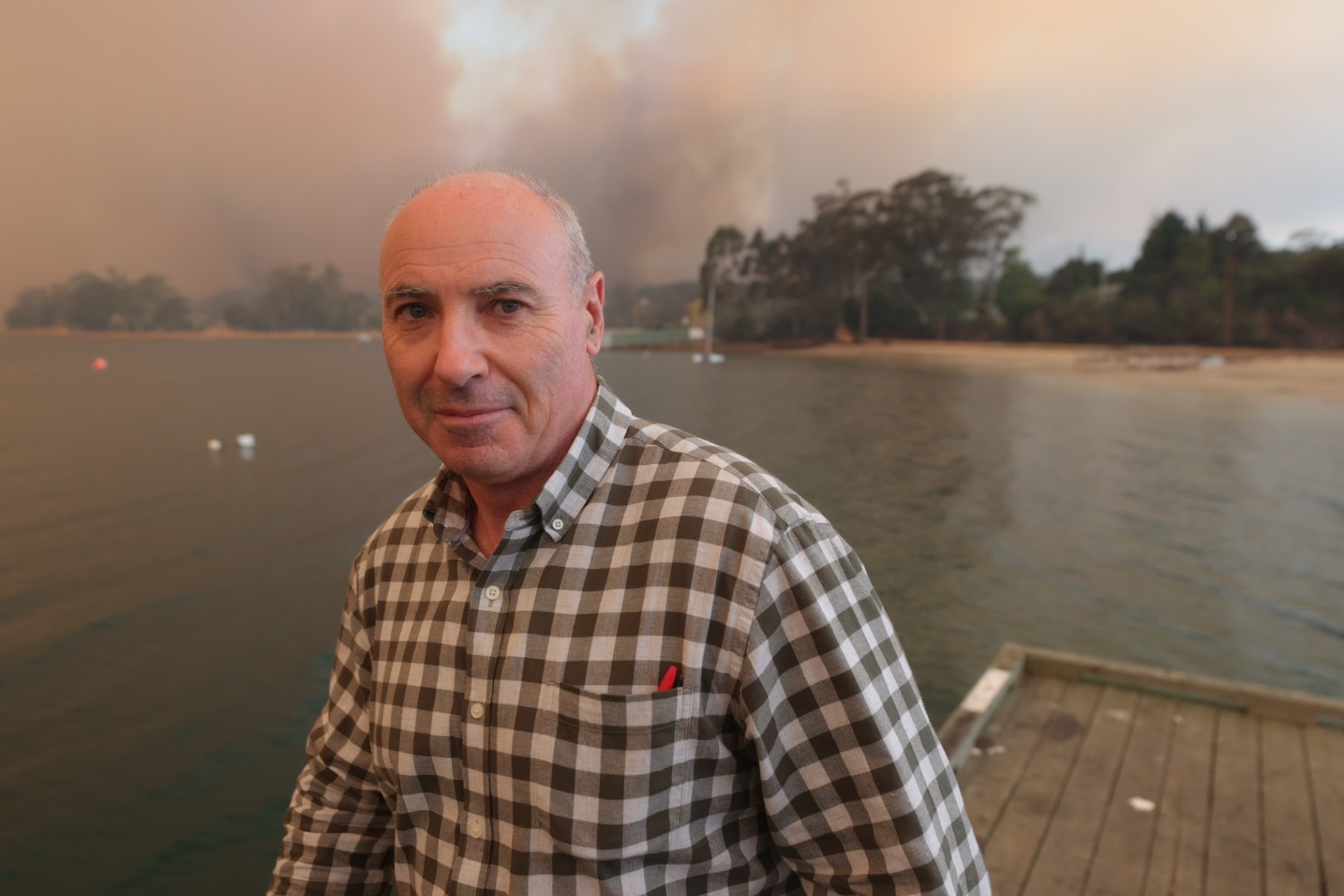 Un hombre con camisa a cuadros se encuentra en un muelle con un incendio forestal ardiendo detrás de él.