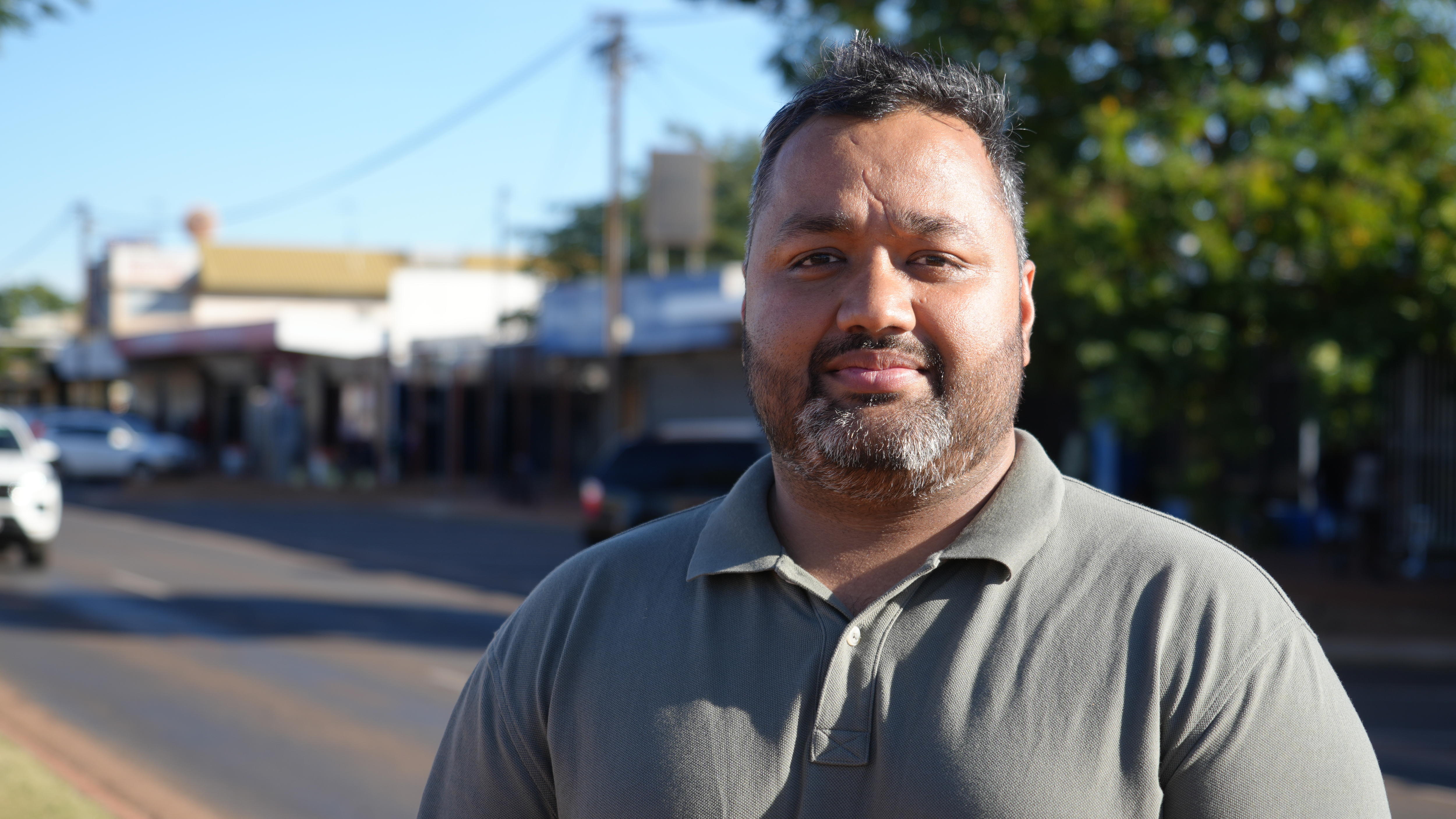 Chest-up shot of Indian-Australian man with black hair and beard, standing street-side with serious expression.