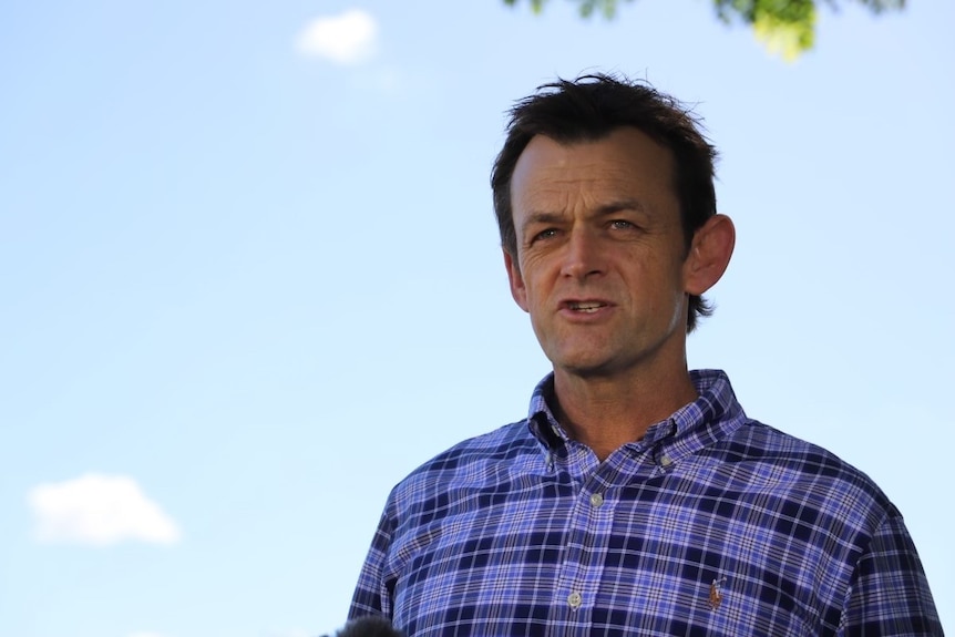 A close-up of a man in a purple and blue checked business shirt looking serious. Blue sky in the background.