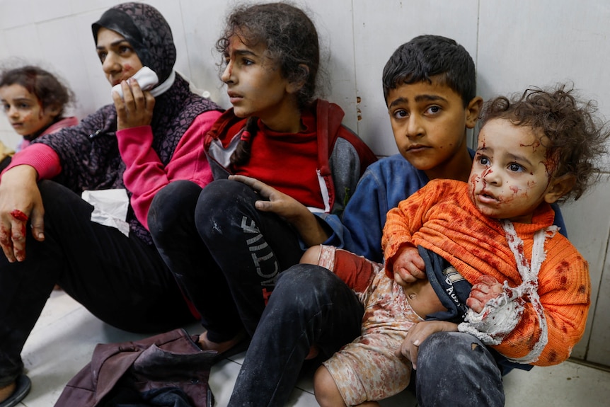 Five kids sit on the floor, dusty and bloodied with one toddler in a sling