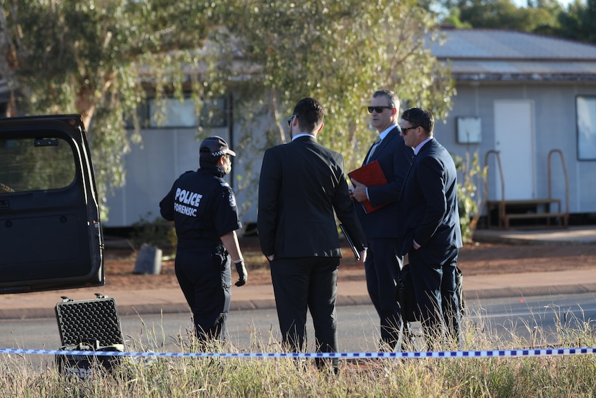 A police forensic officer stands outside with a group of detectives in suits.