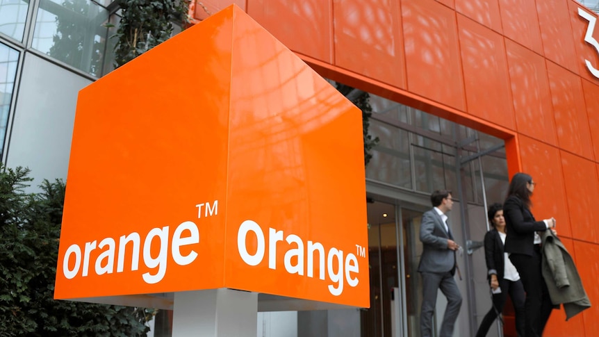 A large square Orange telecom sign is seen in the foreground as three people in corporate attire walk out of sliding glass doors