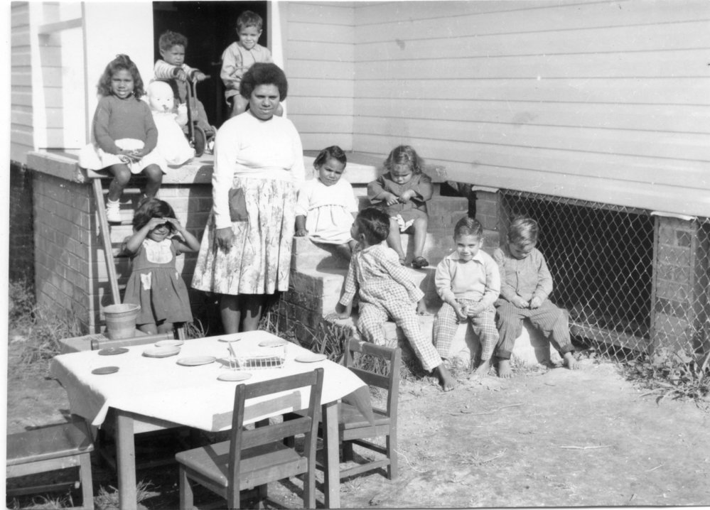 Black and white image of an Aboriginal woman with nine children under 6 sitting on steps, table and chairs outside