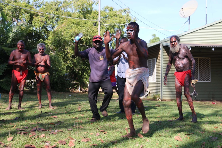 Indigenous men in traditional garments perform a ceremony dance in the yard of a home.