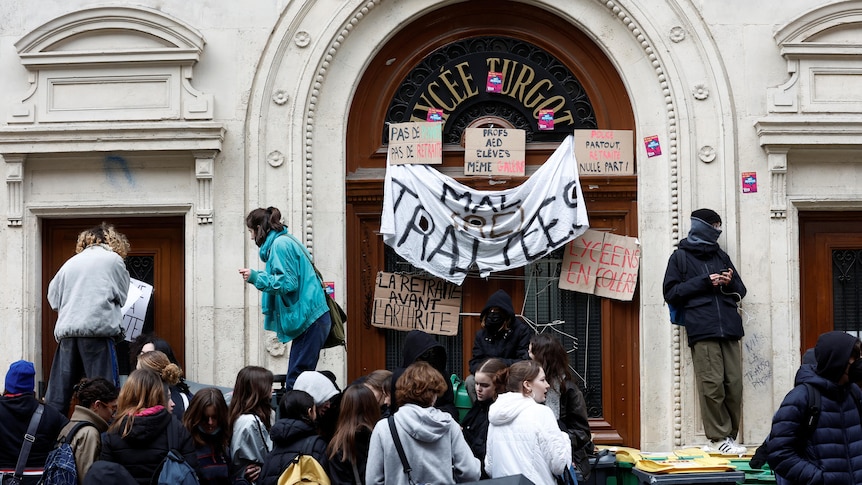 A crowd of young people in winter clothing, some standing on rubbish bins, hang banners and signs over an ornate front door.