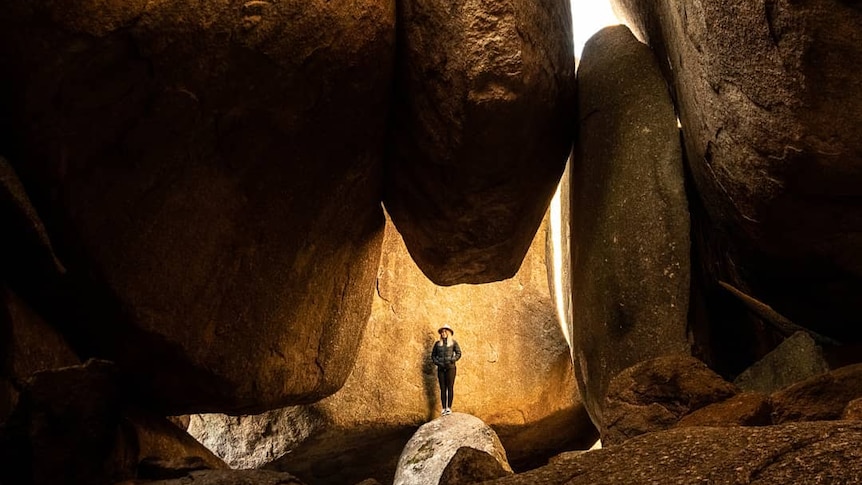 A person stands at the entrance to a cave as sunlight pours in from behind.