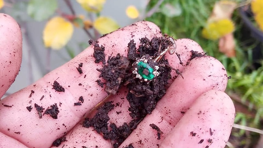 A hand is covered in dirt and holding and emerald and diamond gold ring. In the background is the garden bed. 