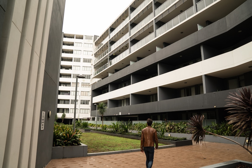 a man strolls along a paved pathway with apartment buildings towering around him.