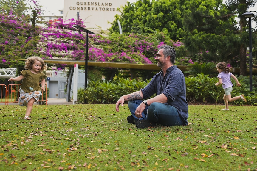 Two little girls run around their dad who's sitting on the grass.