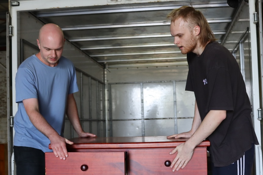 Two men moving a chest of draws from the back of a truck inside a storage shed