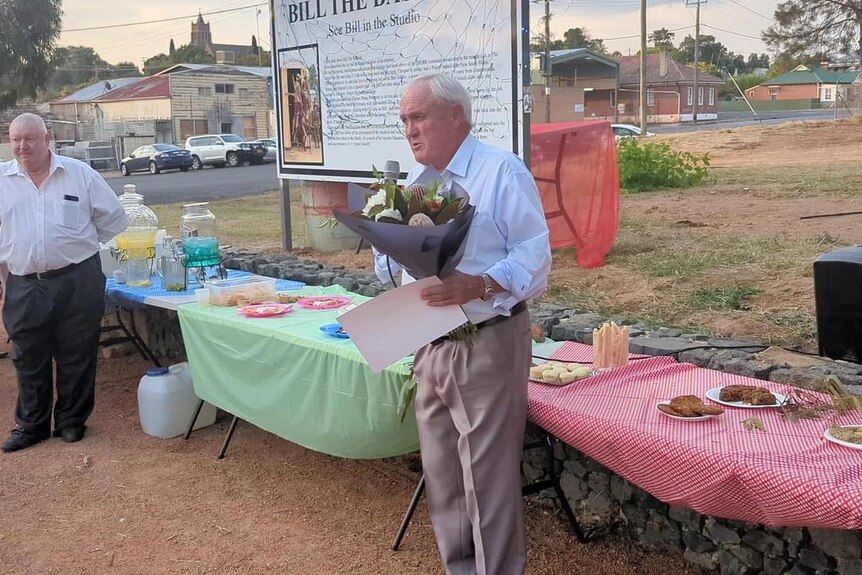 An older man holds a bunch of flowers as he speaks at a community event.