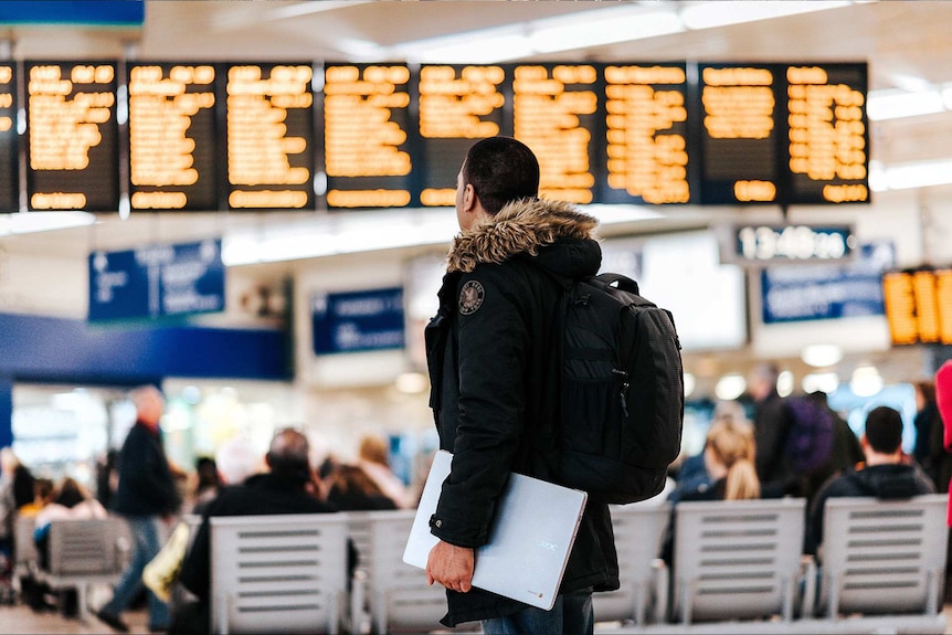 Man wearing a backpack and standing in an airport looking at the departures sign