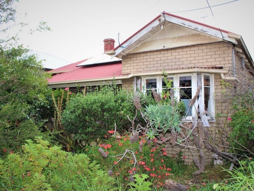 A rundown, cream fake brick facade house with an overgrown garden in suburban Melbourne