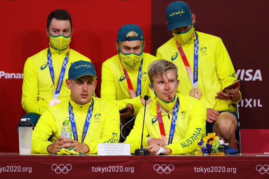 Australian cyclists sit at a press conference - one man has a bandage on his hand and bloody scrapes on his face.