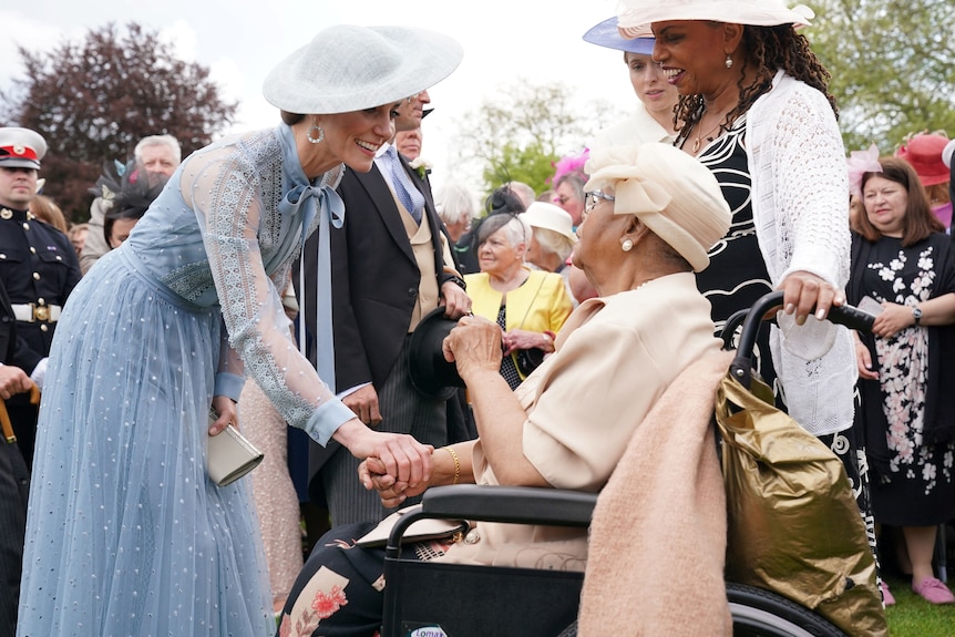 A smiling woman in a blue dress holds the hand of an older woman in a wheelchair who is dressed in beige. 