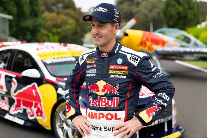 A male supercars driver stands with his hands on his hips in front of his car.