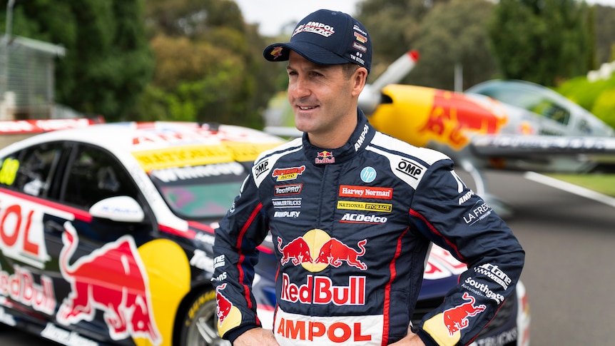 A male supercars driver stands with his hands on his hips in front of his car.