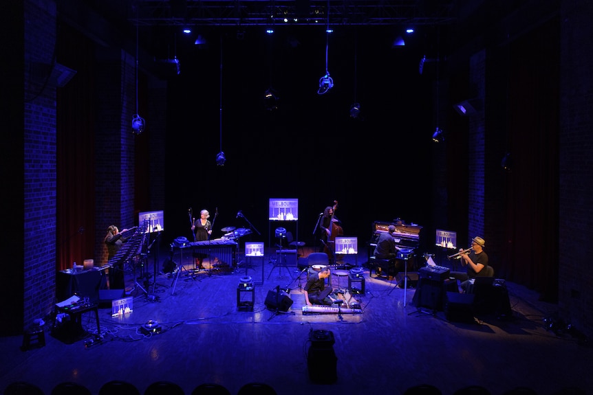 Six musicians play a range of instruments on a blue-lit stage.